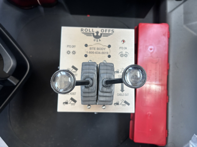 Roll-Offs control panel with two joystick levers labeled PTO OFF, PTO ON, CABLE OUT, and BOOM/HOIST, and a red safety cover; logo and contact info on panel.