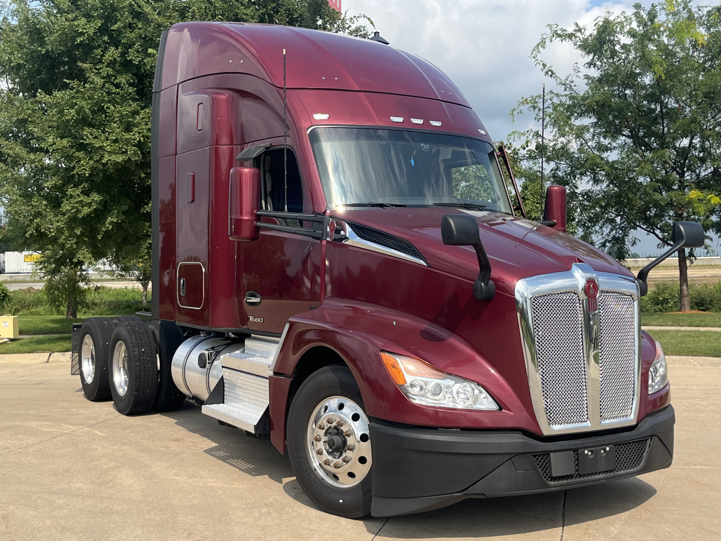 Kenworth T680 tractor in maroon color, front-left view showing large chrome grille, dual rear axles, sleeper cab, and side mirrors.