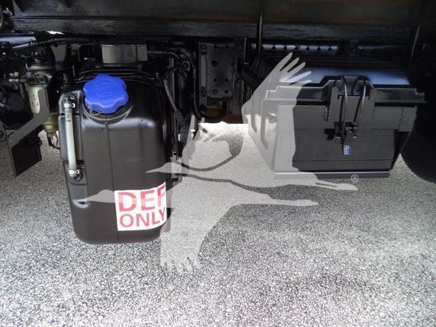 Isuzu NPR-HD undercarriage showing blue radiator cap, coolant reservoir, and large black battery box; DEF only label visible on front of reservoir.