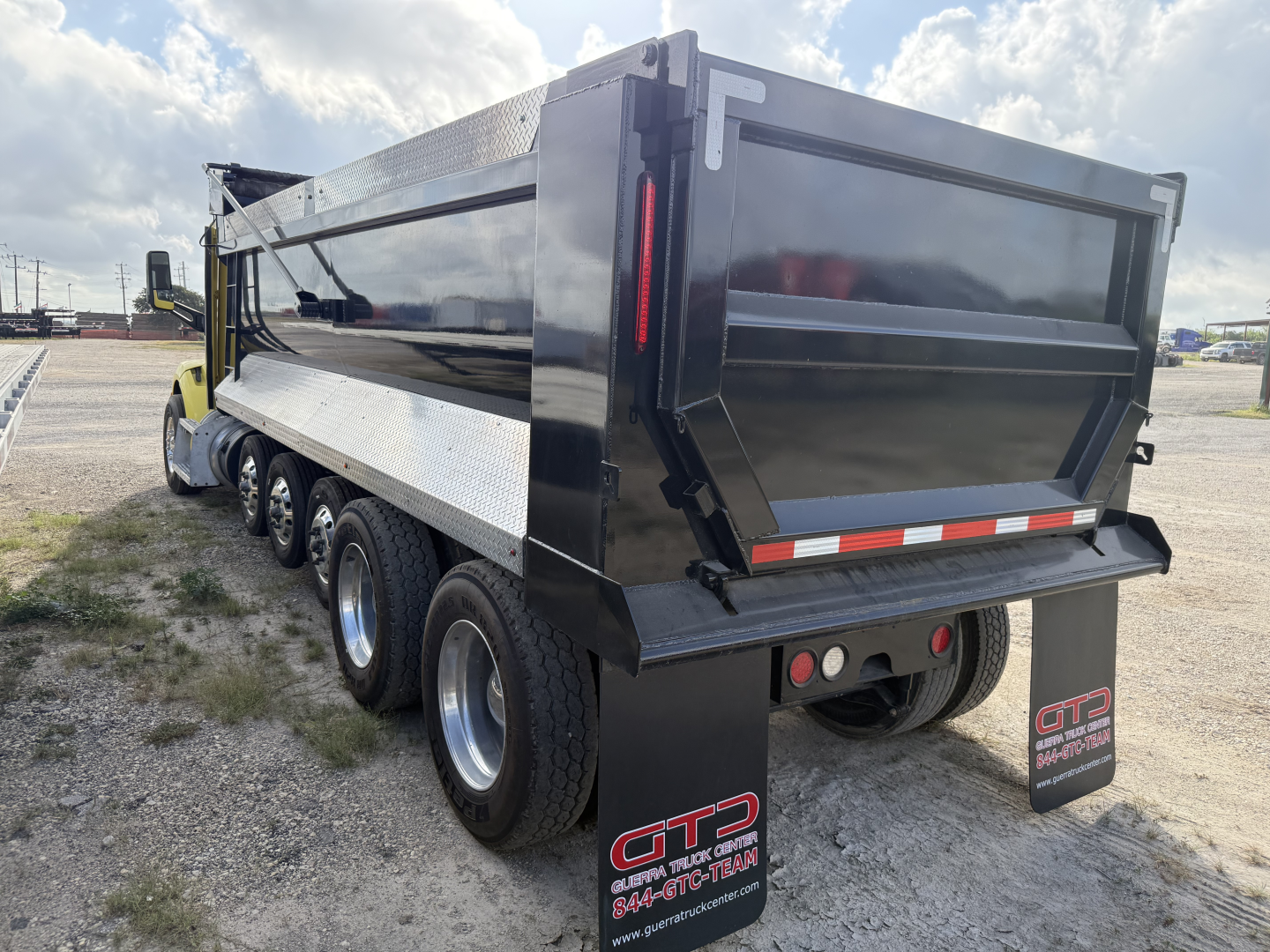 Peterbilt 579 dump truck rear/side view showing dump bed, dual rear axles, black tailgate, mud flaps, reflective strip, and GTD Truck Group sticker.