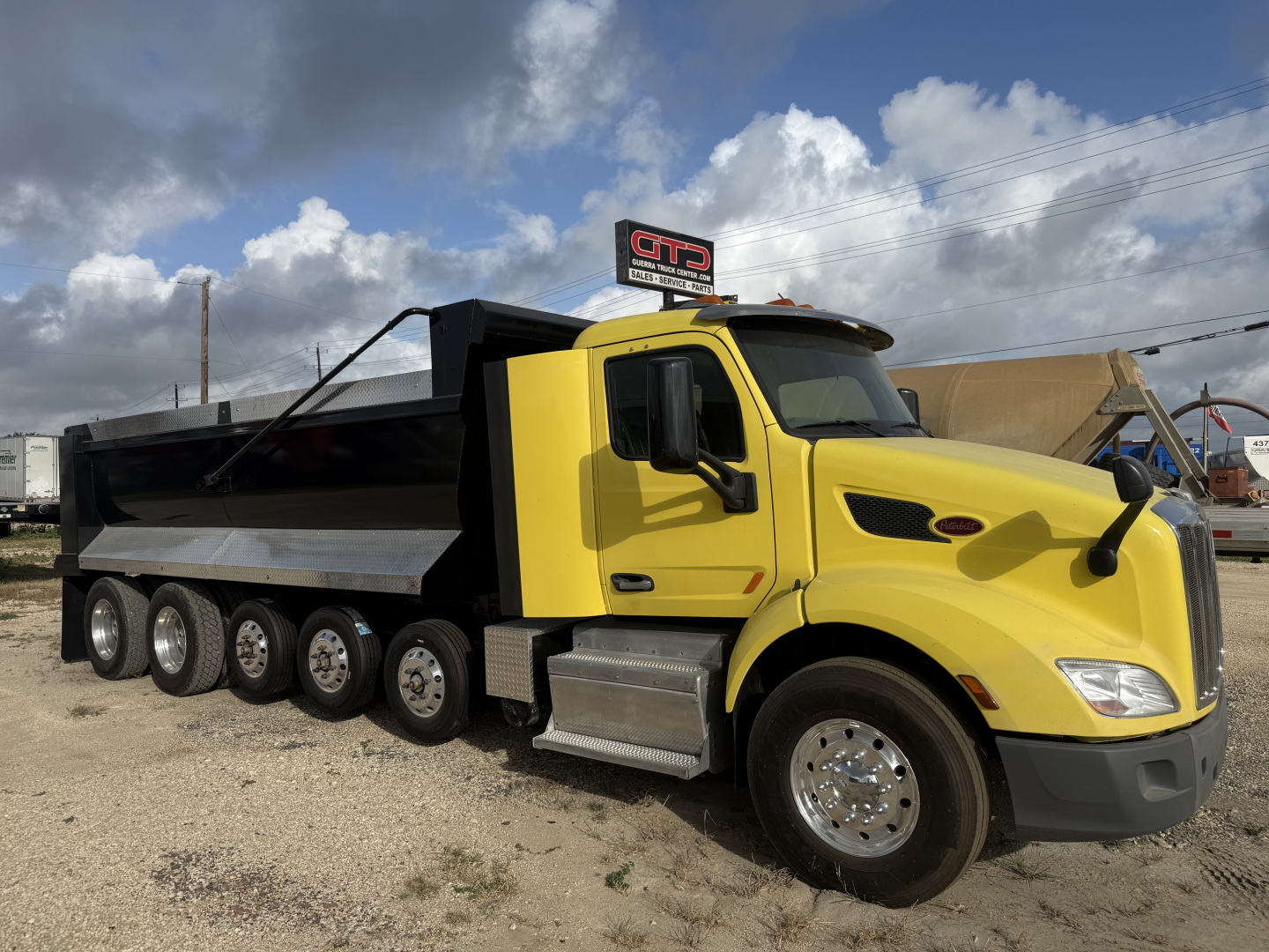 Yellow Peterbilt 579 dump truck with black dump bed, black wheels, and chrome steps. VIN plate visible? Large truck with triple-axle rear. Clear sky.