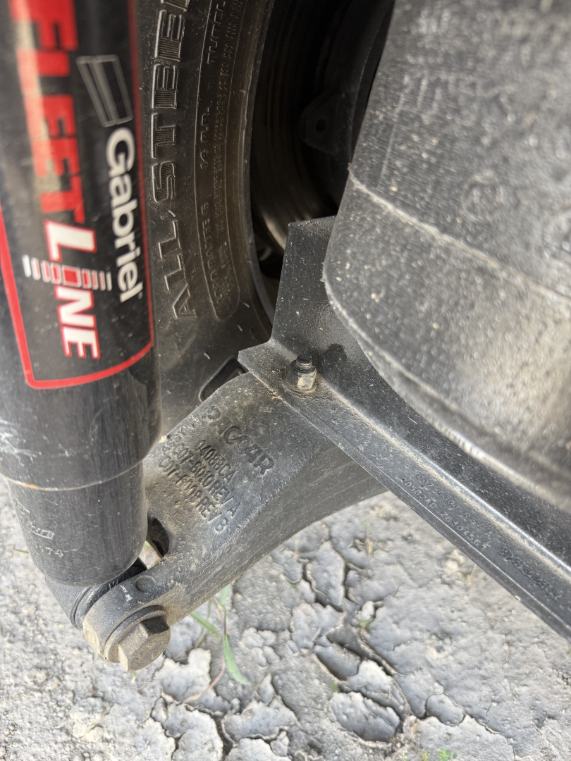 Peterbilt front suspension near wheel shows a Gazelle PureAir shock label and a front leaf spring mount with bolts. Visible text on component includes 'GAZELLE' branding on shock and markings on hub area.