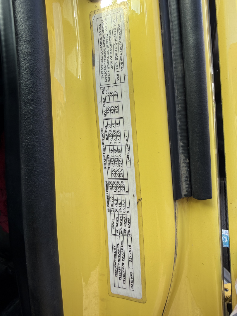 Diesel truck body side with vertical sticker containing vehicle specs and tire/weight data on yellow Peterbilt 579 cab, near door jamb.