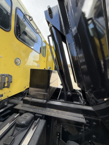 Truck chassis and dump body mounting area with yellow cab visible on left and black hydraulic mechanism in foreground; VIN and model info not visible on image