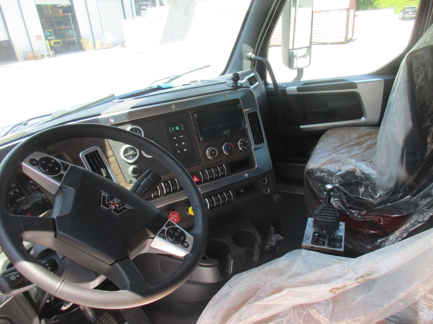 Driver cockpit of a 2027 Western Star 49X dump truck showing steering wheel, dashboard controls, and seating area with plastic covers.