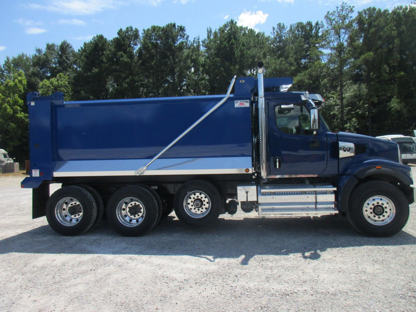 Blue Western Star dump truck in profile view showing side of dump bed, chrome steps, and three rear axles.