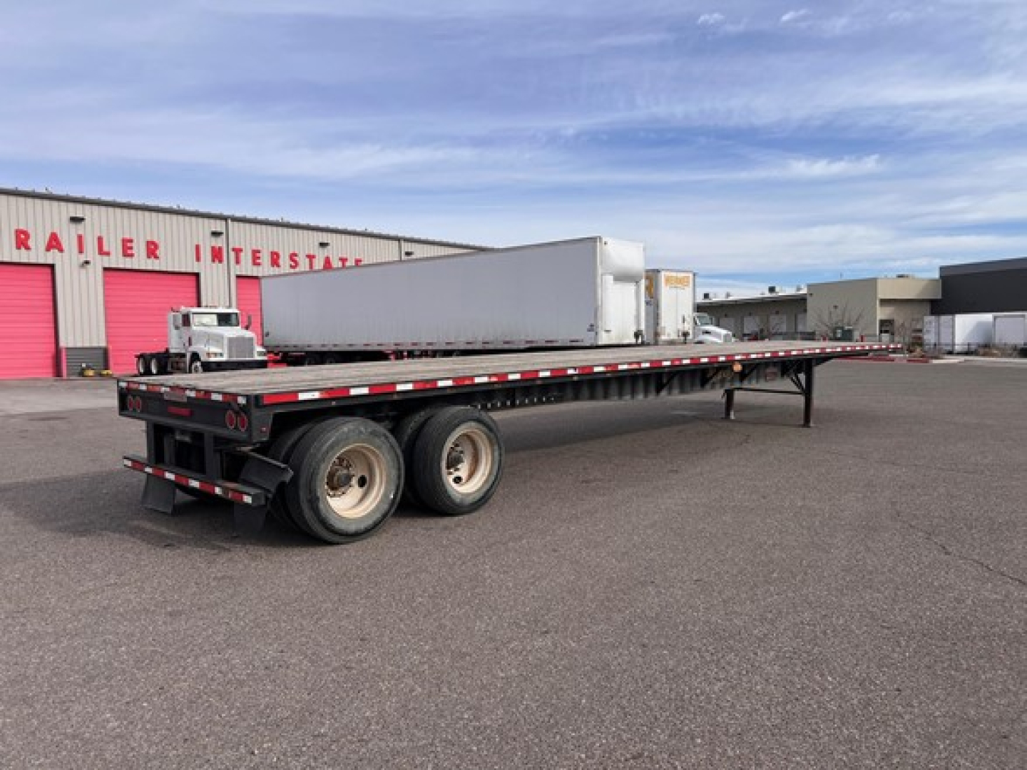 Flatbed trailer showing tandem axles and steel deck with slider, open road in background.