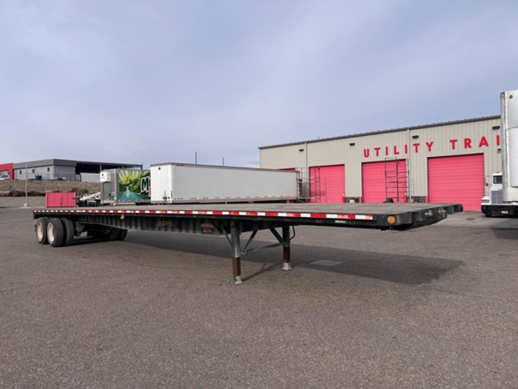 Flatbed trailer in yard showing long steel deck, tandem axles at rear, jack stand near front, with red/white safety rails and a white toolbox. Fontaine closed tandem w slider steel flatbed trailer.