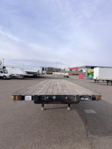 2014 Fontaine closed tandem w slider steel flatbed trailer viewed from front, showing wooden deck, undercarriage, and stabilizing legs.
