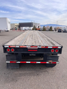 Flatbed trailer viewed from the rear showing wooden deck with tandem axles and black rear bumper, red taillights, and reflective strips.