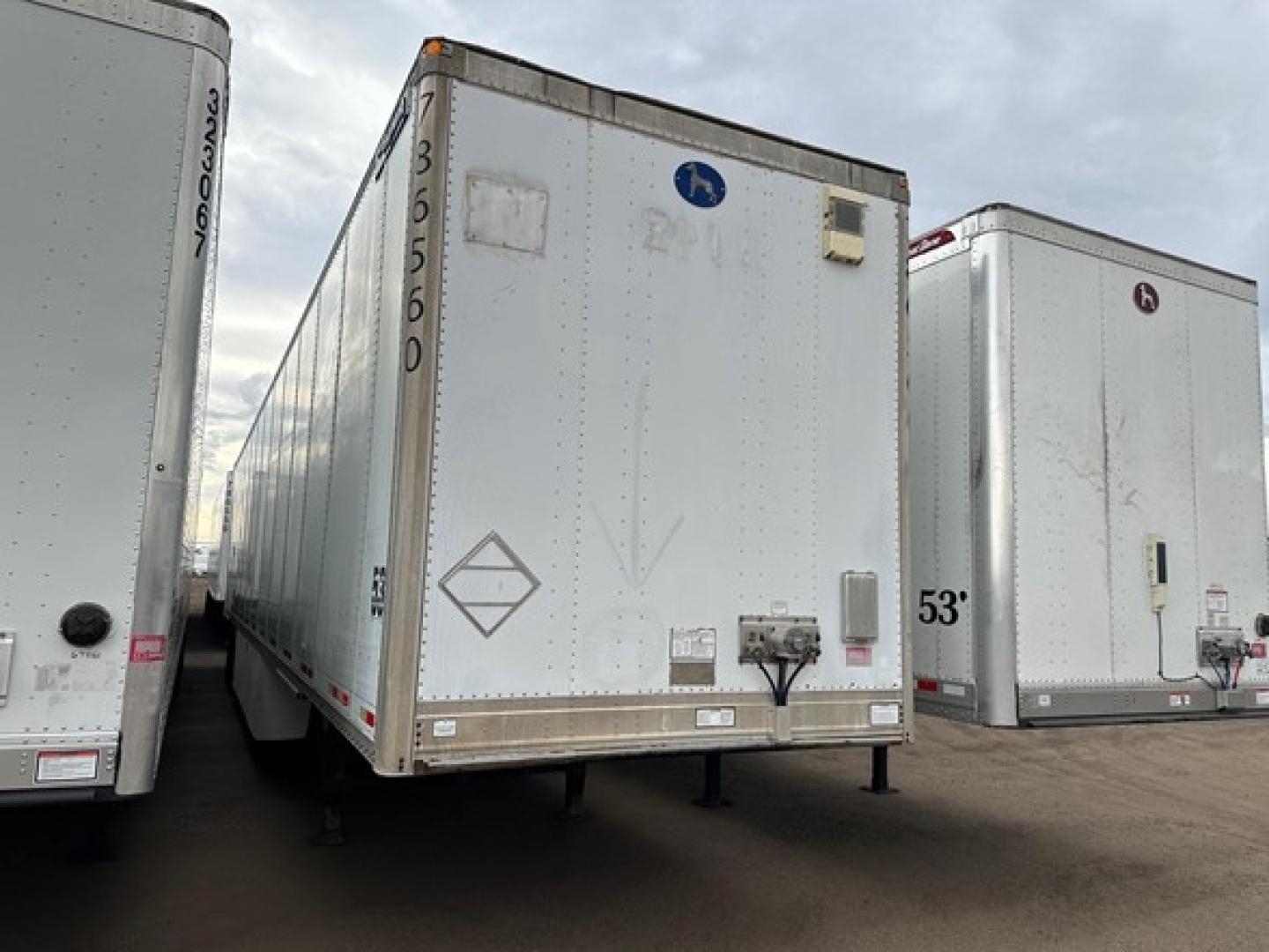Trailer showing rear view of a white Great Dane Champion trailer with a raised rear door, side markings, and a blue circular logo; side of another trailer visible on left in a dealership lot.