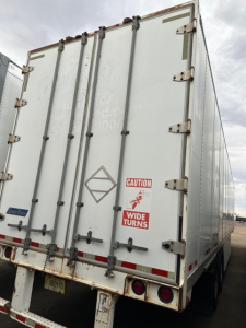 Trailer rear doors with hinges and latches; 'CAUTION WIDE TURNS' sticker; diamond placard outline; tail lights and license plate area visible.