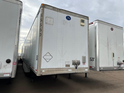 Trailer showing rear view of a white Great Dane Champion trailer with a raised rear door, side markings, and a blue circular logo; side of another trailer visible on left in a dealership lot.
