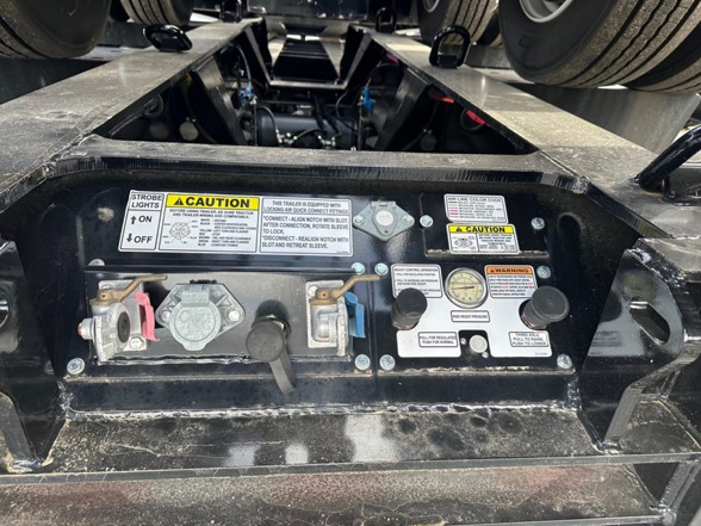 Trailer hitch area with control panel and warning labels on Load King lowboy trailer showing caution stickers, valve levers, and gauges.