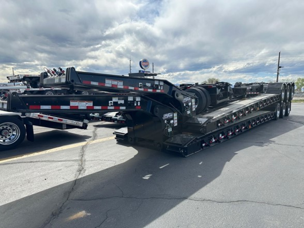Lowboy trailer showing multi-axle transport deck with hydraulic gooseneck and side rails, black with reflective tape; loads of equipment visible in background.