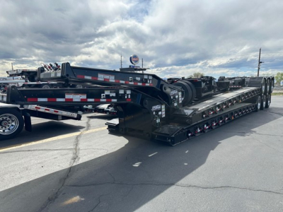 Lowboy trailer showing multi-axle transport deck with hydraulic gooseneck and side rails, black with reflective tape; loads of equipment visible in background.