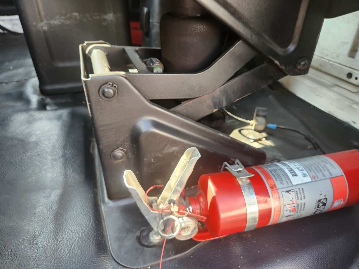 Fire extinguisher mounted in truck bed near metal bracket and wiring, with label text on extinguisher visible.