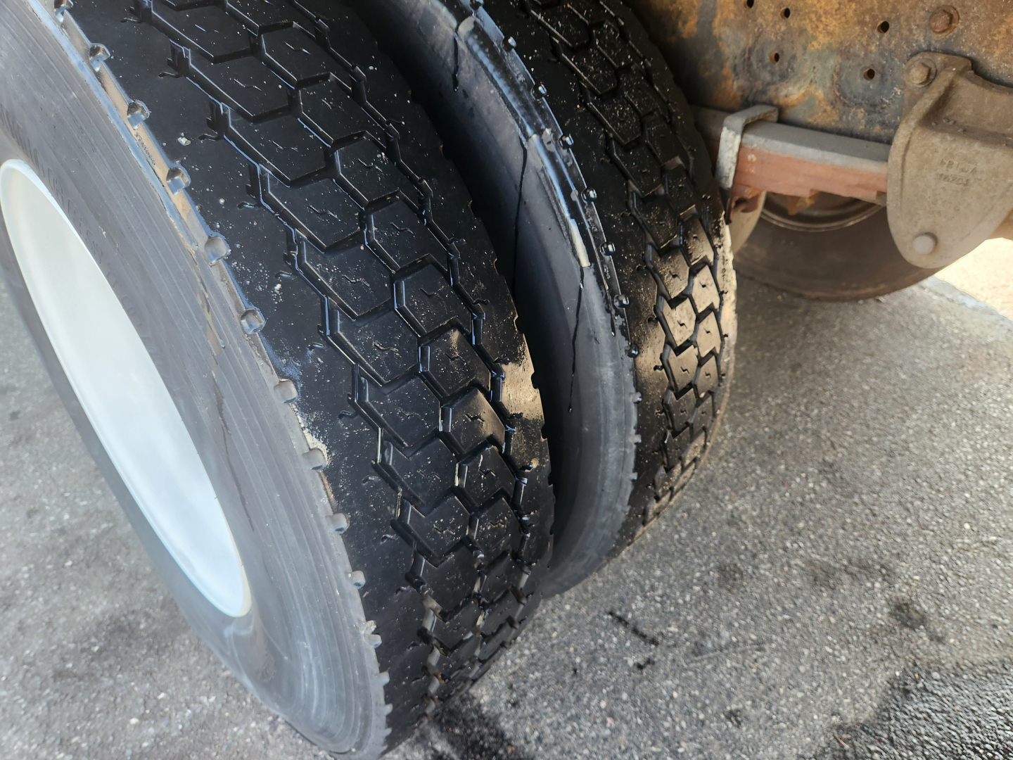 Truck wheel and dual tire setup showing tread pattern and inner wheel; part of chassis and undercarriage with some rust-colored components.