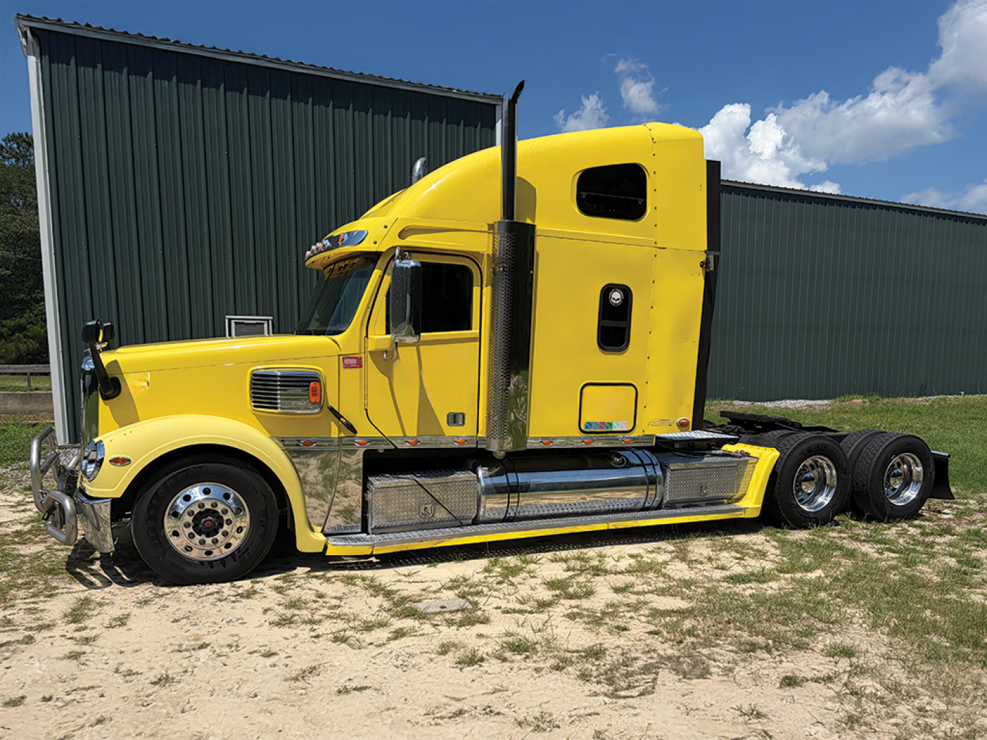 Yellow Freightliner Coronado truck tractor side view showing sleeper cab, chrome exhaust stacks, fuel tanks, and 3-axle rear setup.