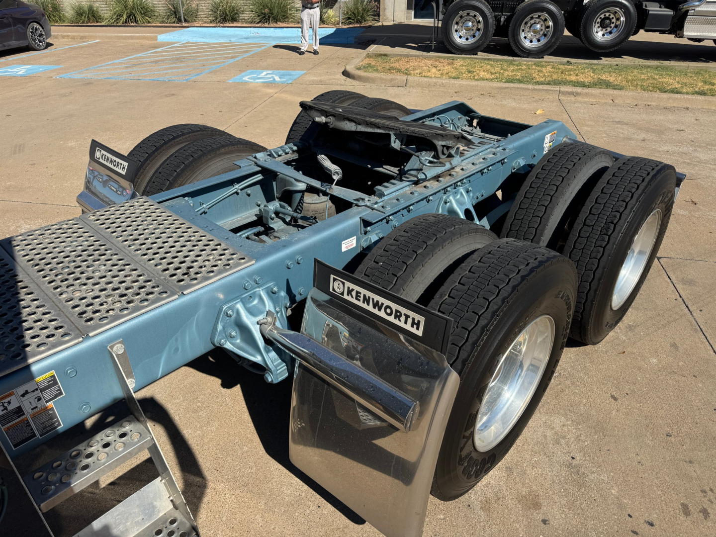 Kenworth chassis with dual rear axles and Kenworth mud flaps; blue frame showing wheel rims and platform; no cab attached; truck yard in background.