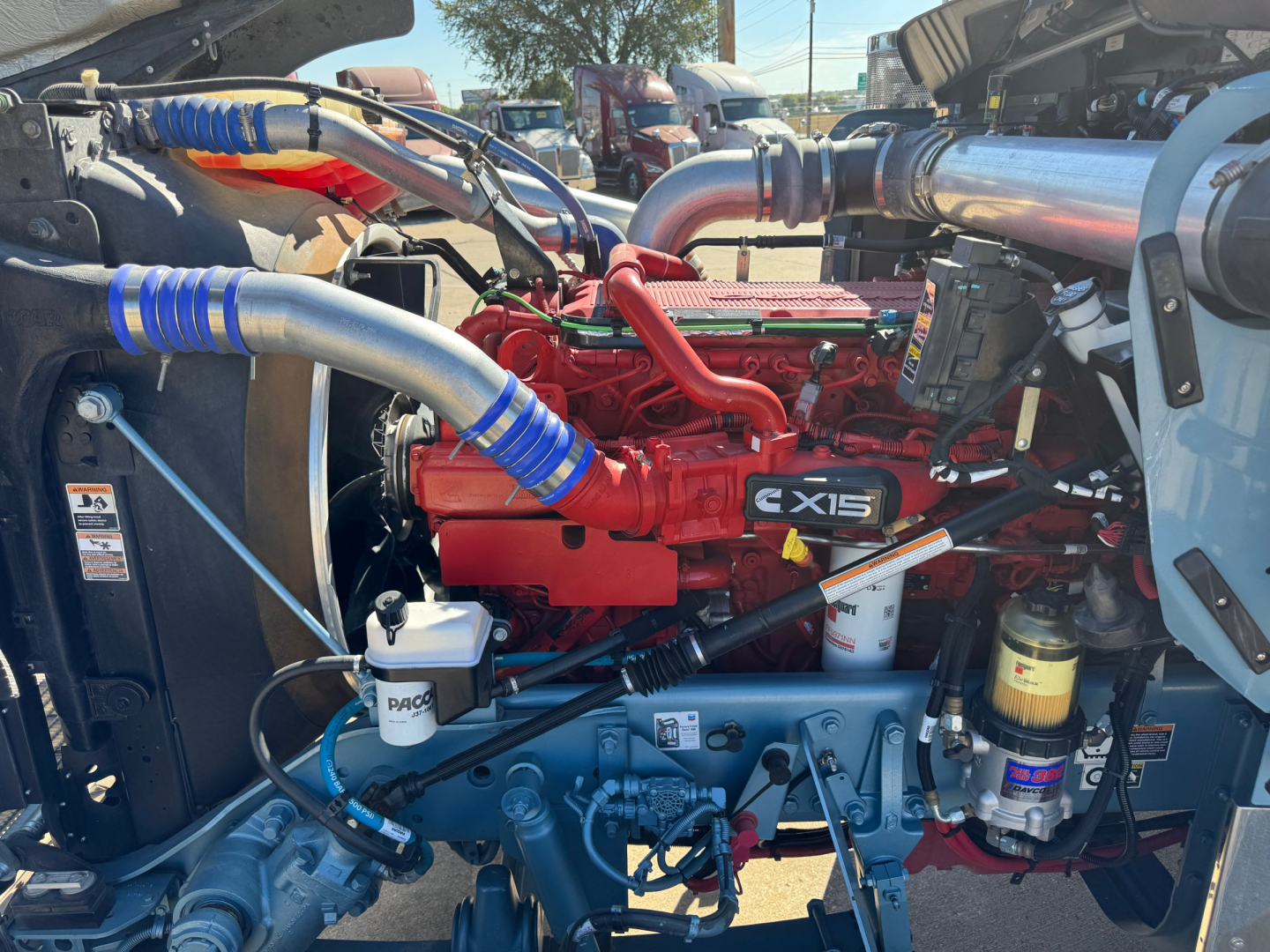 Kenworth W900L engine bay showing red engine with blue hoses, silver intake pipe, black radiator, and various filters. Visible labels include CXS15 on engine and generic oil/air filter components.