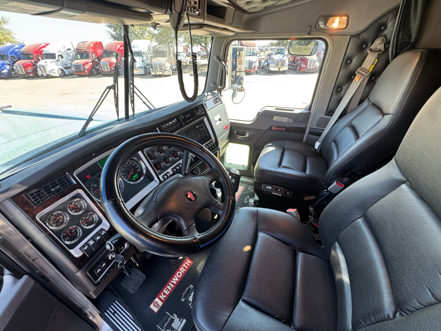 Kenworth W900L truck interior showing steering wheel, dashboard with gauges, driver seat and passenger seat in a sleep cab; visible outside are other trucks in a dealership lot.