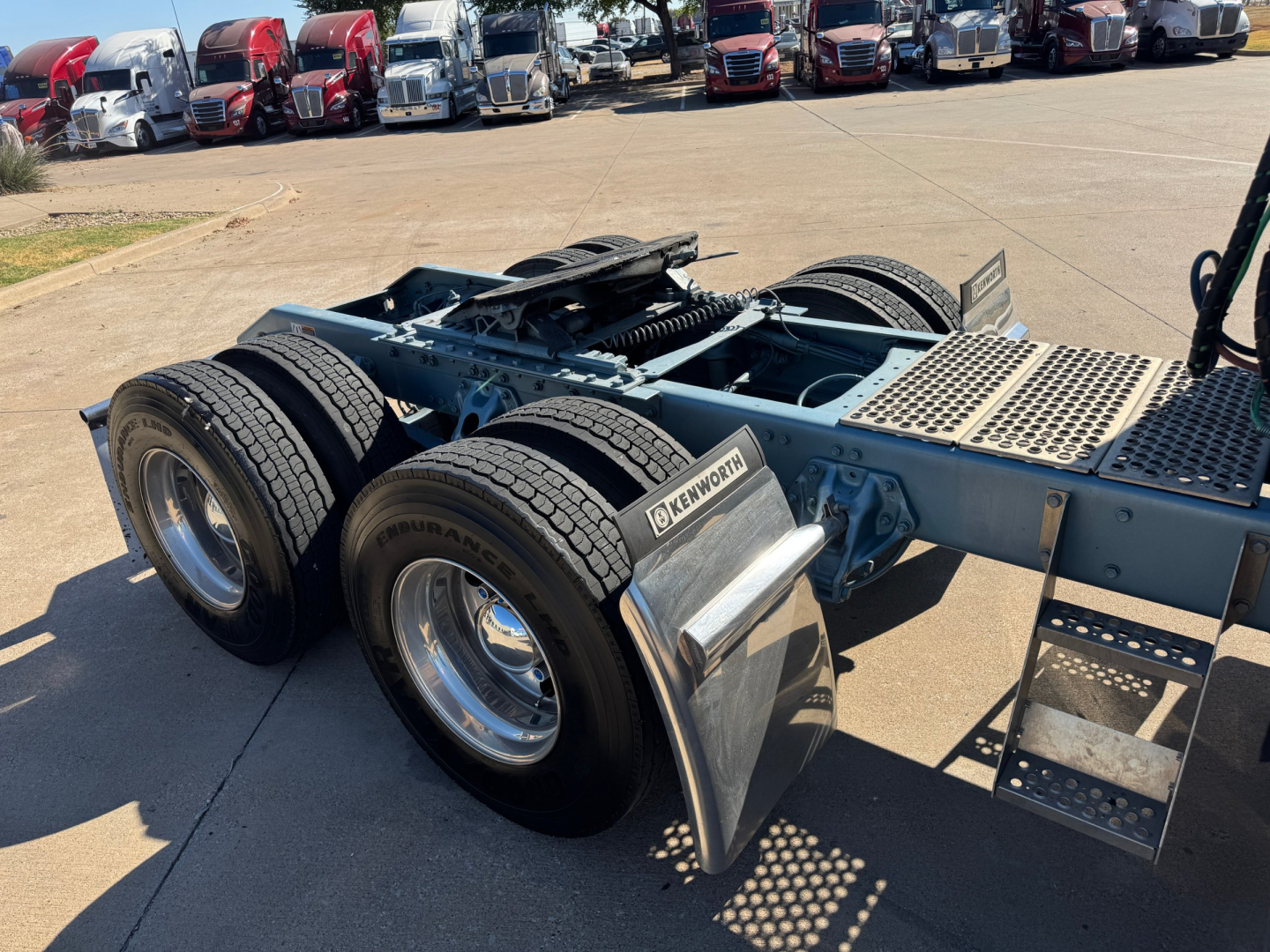 Kenworth W900L tractor chassis with dual rear axles and blue frame, metal steps, mud flaps, and cabless rear section visible in a truck lot.