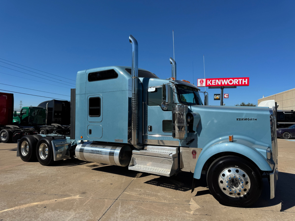 Kenworth W900L conventional sleeper truck in light blue, side view showing chrome exhaust stacks, fuel tanks, and steps.