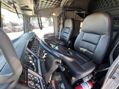 Truck cab interior of a 2025 Kenworth W900L showing driver seat, steering wheel, dashboard, and control panels.