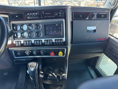 Kenworth W900L dashboard with gauges, infotainment screen, climate controls, and gauge cluster; steering column left; gear shifter visible; center console and cup holders.
