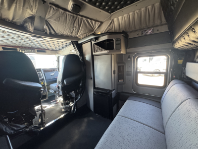 Kenworth W900L truck cab interior with driver seats, passenger seating along window, overhead storage, and a small refrigerator area visible.