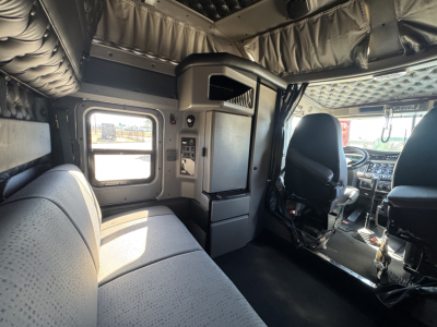 Kenworth W900L interior cab view showing sleeper area and driver seat with instrumentation, black dashboard, and overhead padding.