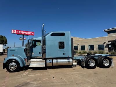 Kenworth W900L truck in blue with sleeper cab, side view showing chassis and wheels, dealership lot in background.