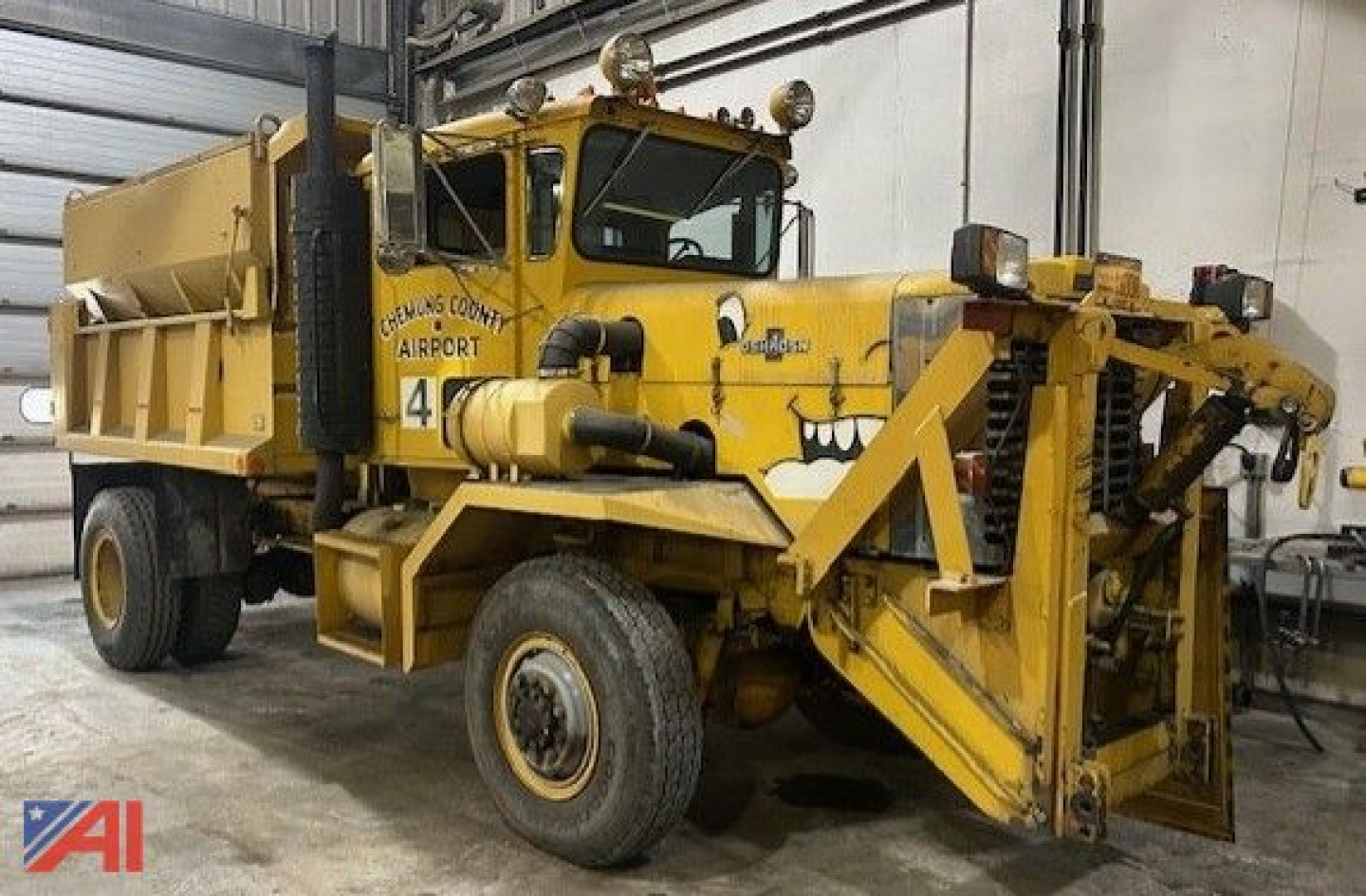 Front-left view of a yellow Oshkosh P2323-6 dump truck with a sander attachment inside a workshop. Visible markings include Oshkosh County Airport, a number 4, and various mechanical components.