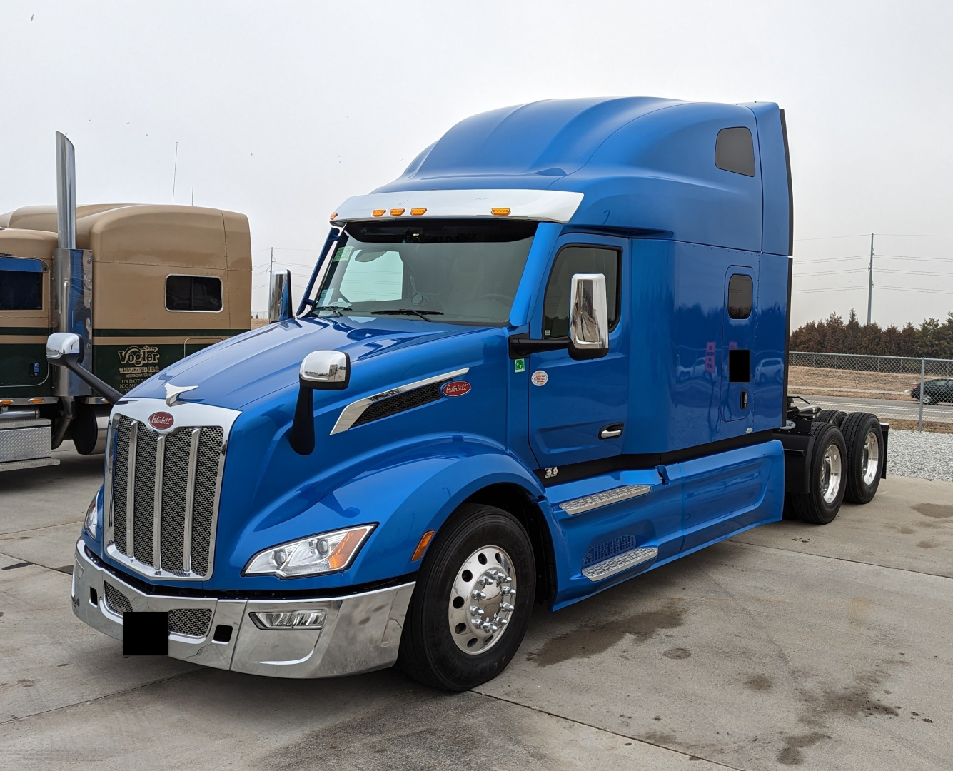 Peterbilt 579 conventional sleeper truck in blue, front left side view. Visible text includes Peterbilt emblem on grille, chrome bumper, and small badge on door. No readable VIN or numbers clearly visible.