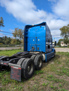 Blue Peterbilt 579 truck cab and chassis side view showing triple rear axles, rear mudflaps with text, and wheel tires.