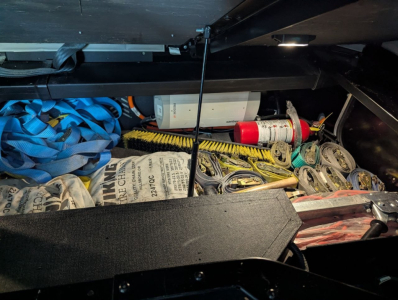 Trunk/storage compartment showing miscellaneous tools and items, including a blue strap, yellow brush, fire extinguisher, and assorted cables.