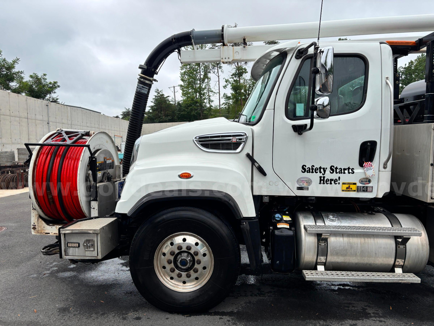 Truck cab side profile showing suction hose reel and warning decals on Freightliner 114 SD with reel at front and hose outlet; front wheel visible