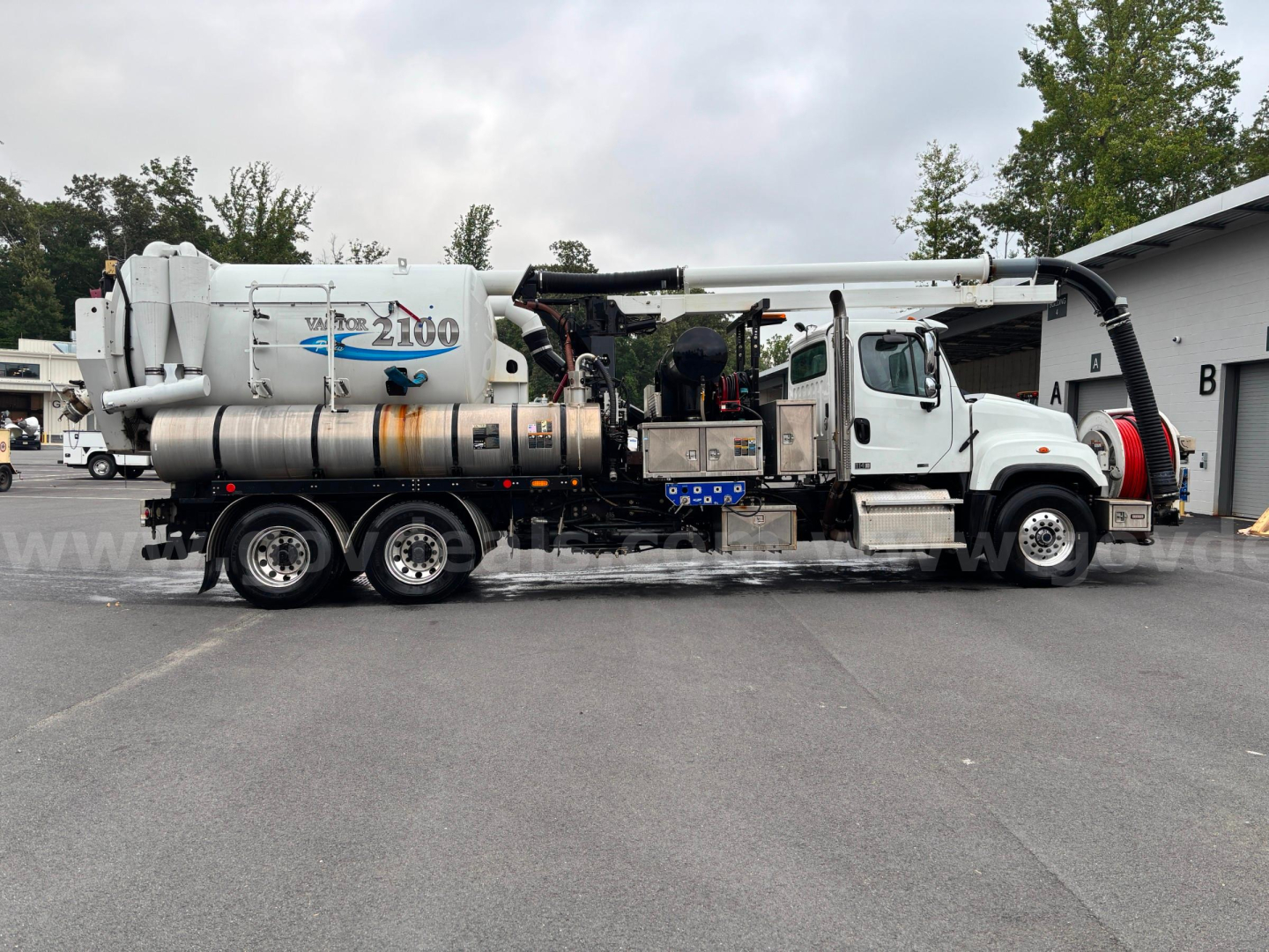 Truck-mounted vacuum tanker side view with large white cylindrical tank and integrated hose reel; visible labels on tank; Freightliner 114 SD utility truck with rear vacuum unit.