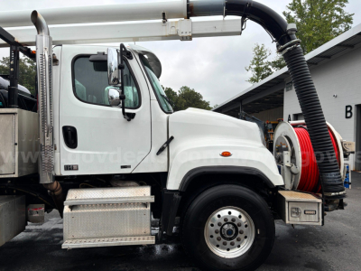 Freightliner vacuum truck cab and hose reel setup with white body and large black vacuum hose on right side.