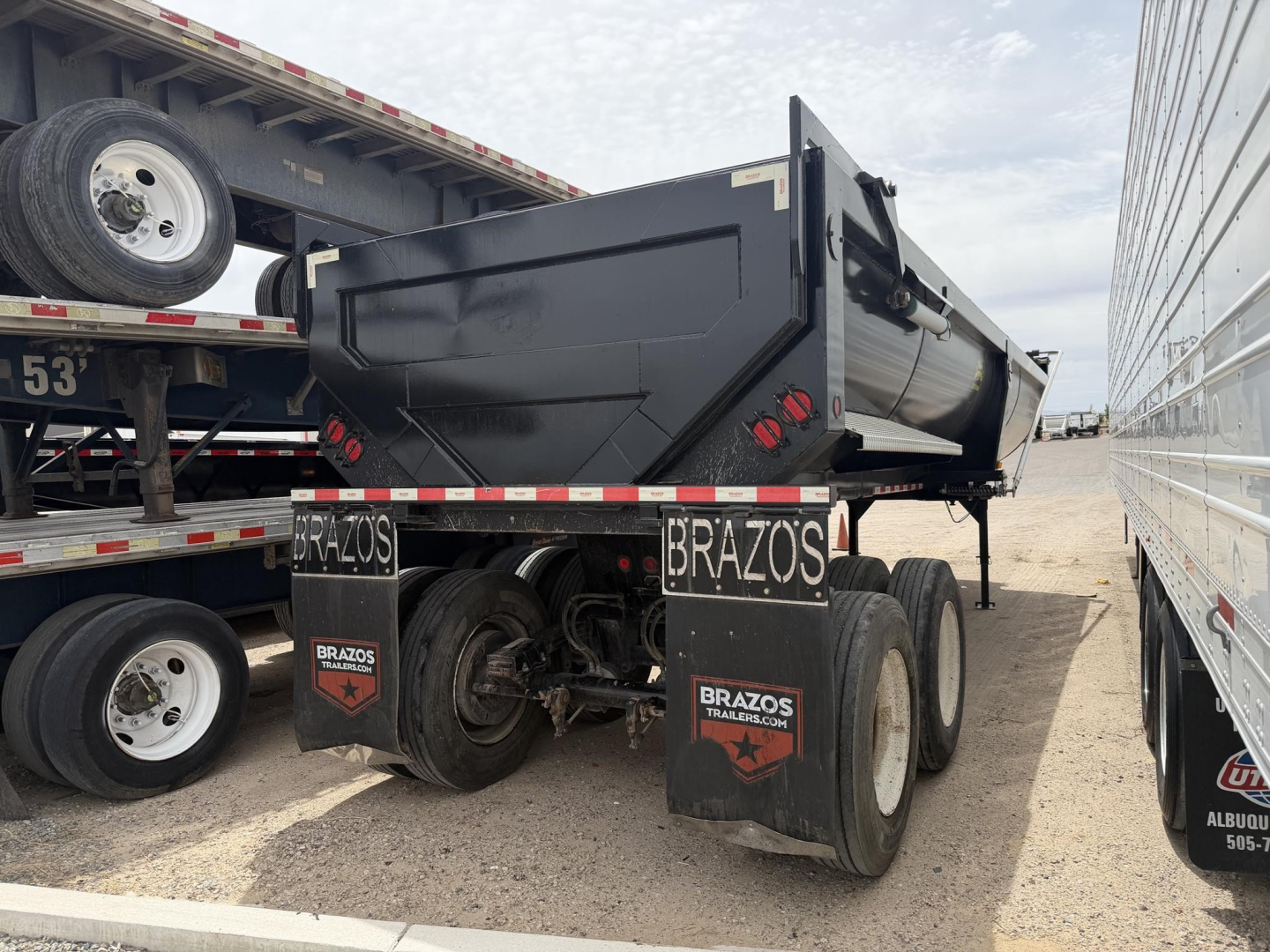 Truck trailer end dump showing Brazo s high-lift gate and frameless steel bed with rear bumper and taillights, black color, storage area, mudflaps with Brazo s branding.