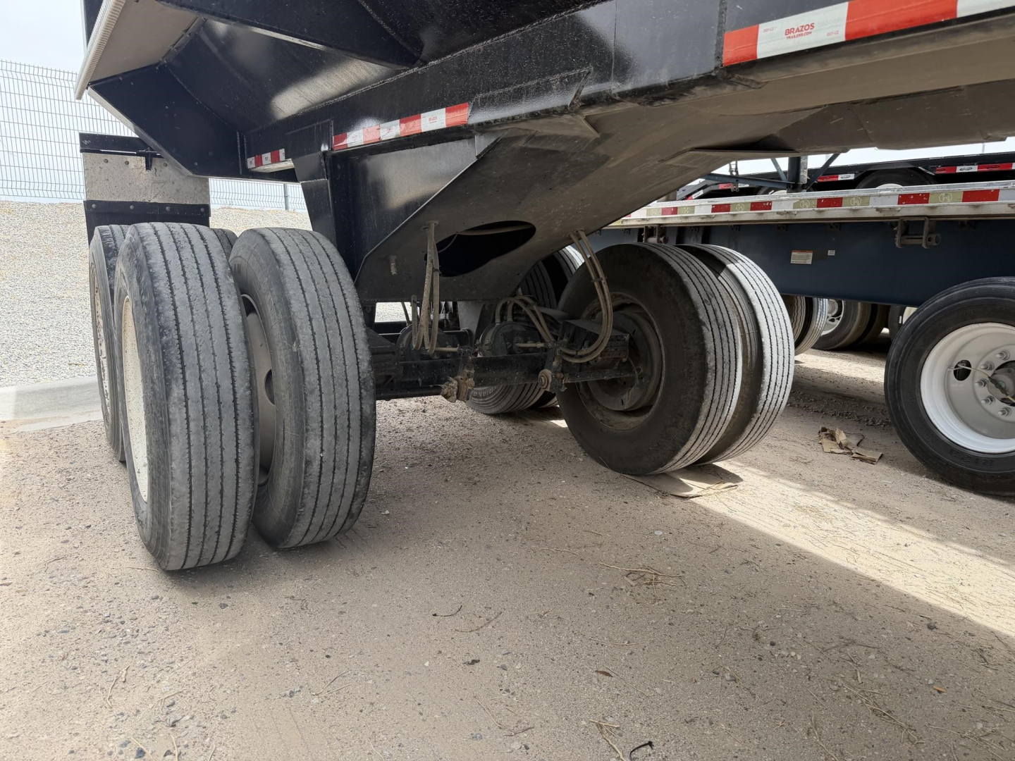 Dump trailer undercarriage showing twin rear axles and exposed wheel hubs with tires; framed steel frame and hydraulic components visible under a frameless rock end dump.
