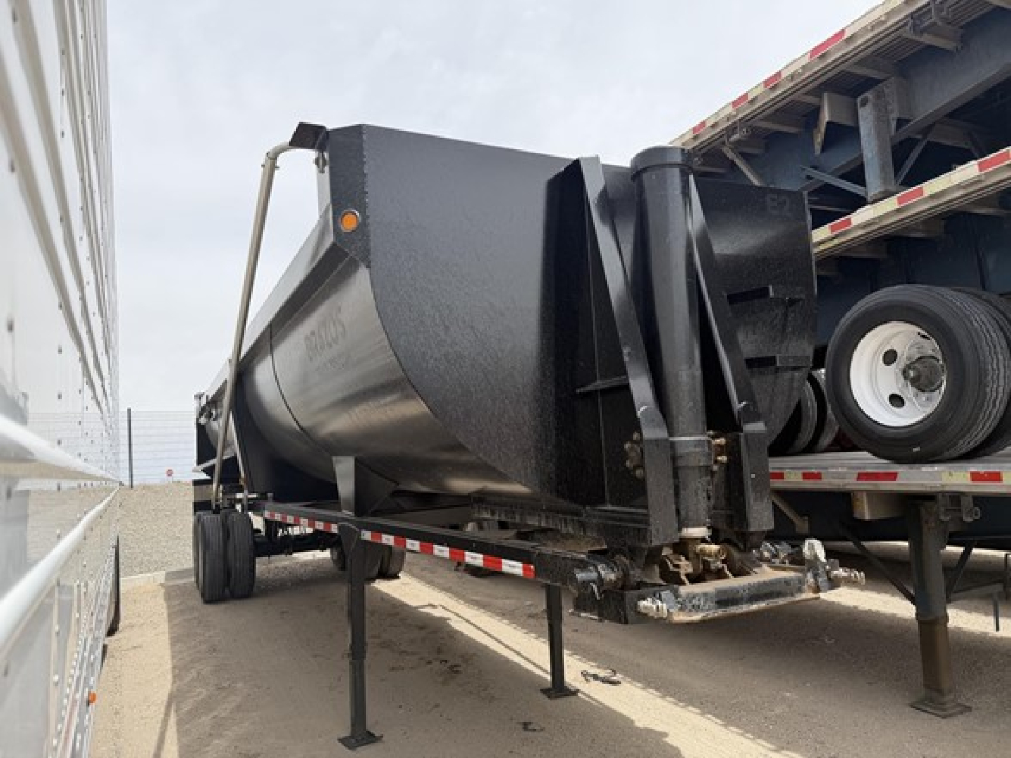 Frameless rock end dump trailer in a yard showing triangular hitch and support legs, side view with ramp and truck bed in background.