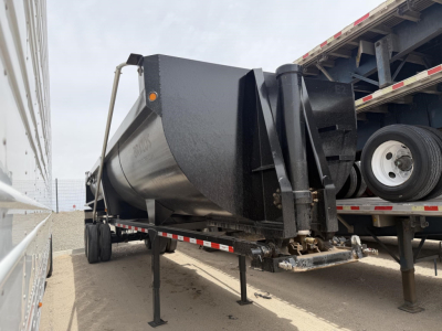 Dump trailer with high lift gate in a yard, steel frameless body, black front end, supporting legs down, attached to a truck, visible wheels and trailer bed.