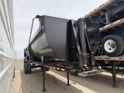 Frameless rock end dump trailer in a yard showing triangular hitch and support legs, side view with ramp and truck bed in background.