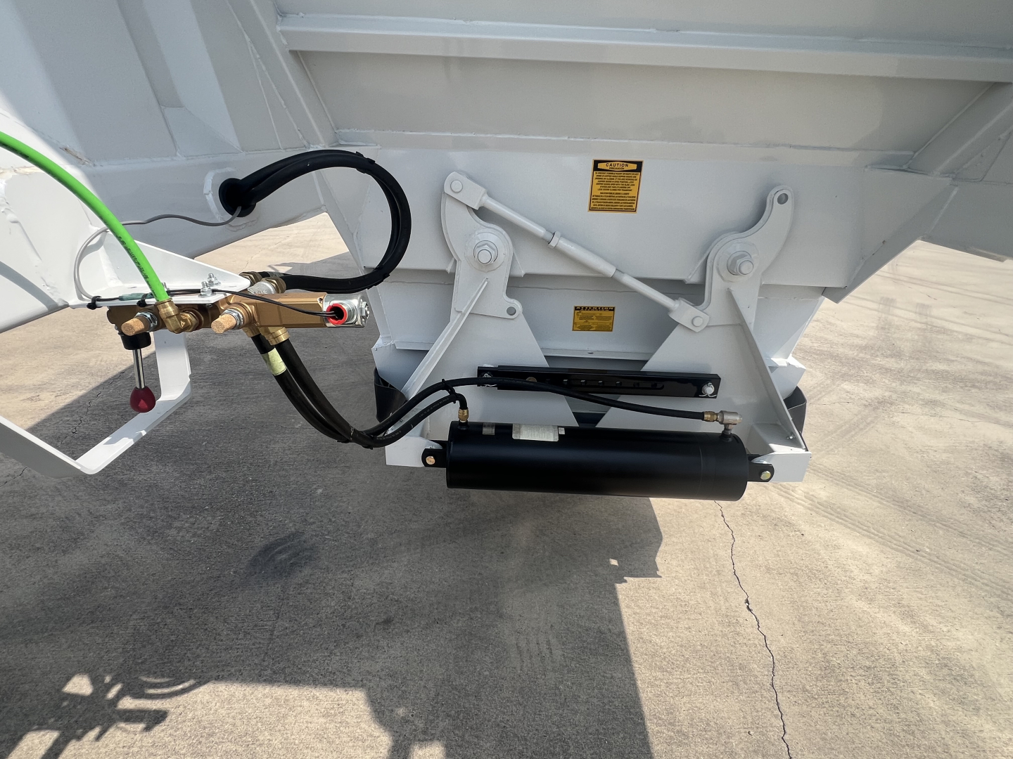 Trailer hitch area showing air ride suspension cylinder and hydraulic lines on a white dump trailer, with yellow warning labels and mounting brackets.