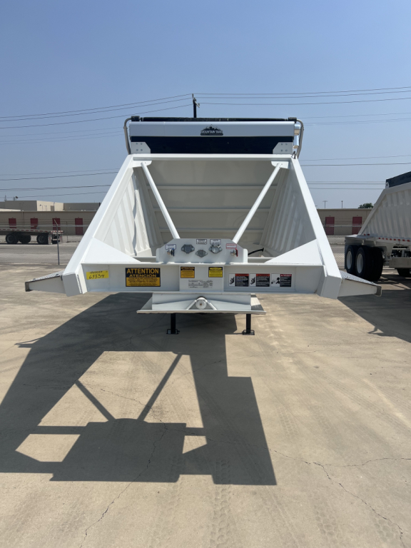 Dump trailer rear view showing white chassis and triangle braces with warning labels and hydraulic components.