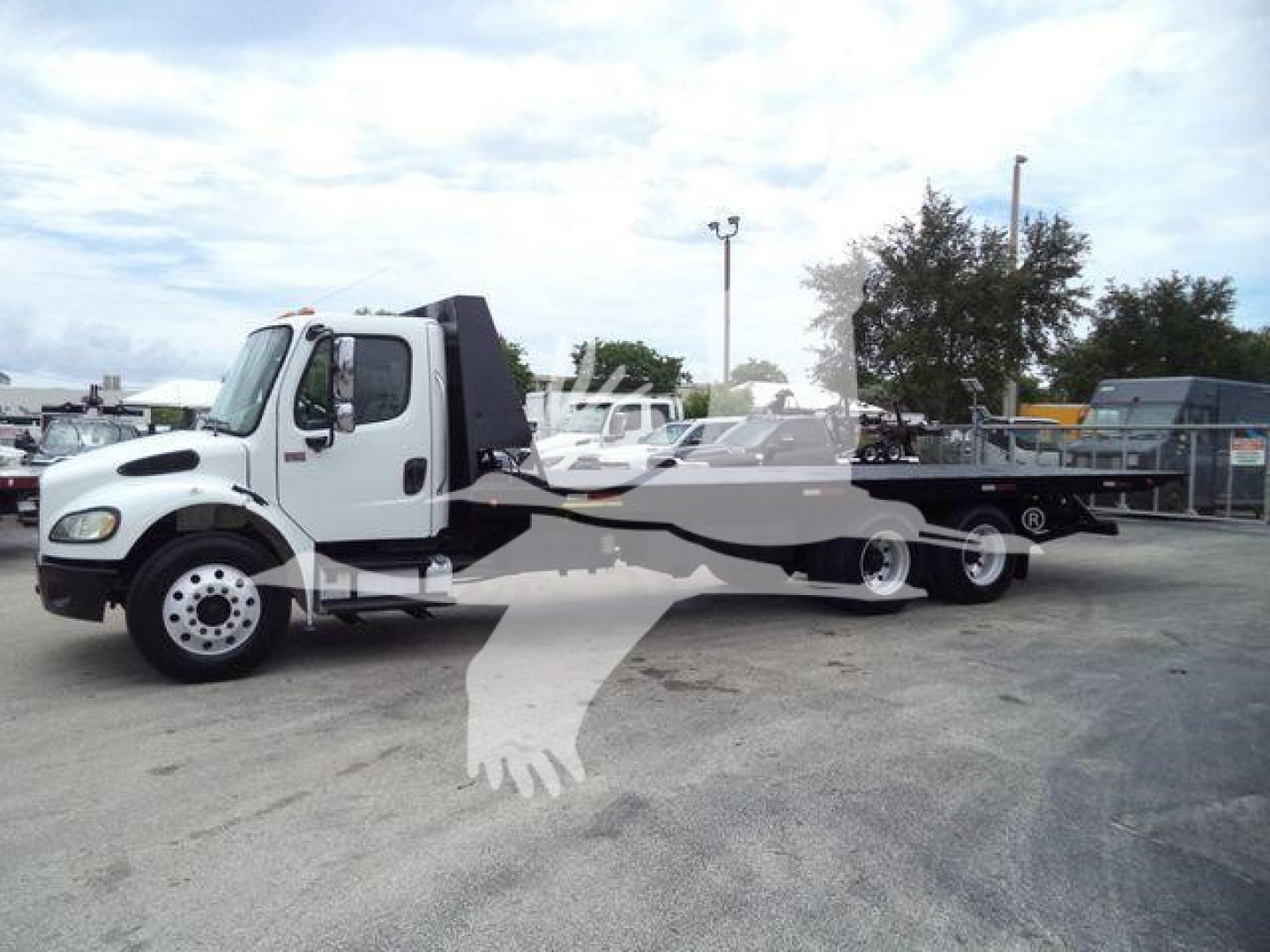 Truck showing a Freightliner M2 106 roll-off towing setup with white cab and long flatbed body, front angle view.