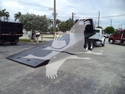 Flatbed style roll-off truck in a parking lot with a large lowered metallic ramp and container pickup behind. Visible text: none legible on vehicle, stock number 2006M2ROLLOFF in listing context.