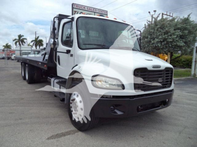 Truck showing white Freightliner M2 106 roll-off bed with dual rear wheels and flatbed area, front view, dealership lot.
